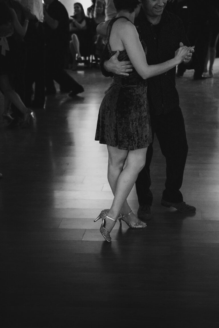 Couple practicing a relaxed two-step and underarm turn on a wooden dance floor.