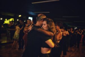 Adults practice partner dancing on a wooden floor while others chat near the edge of the room.