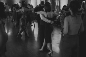 Beginners practicing simple country dance steps on a wooden floor while other dancers move nearby.